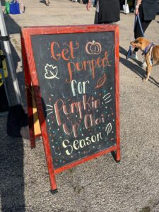 Our updated chalkboard sign at Lincoln Park farmers market with a festive fall design announcing the return of Pumpkin Up Chai. The sign reads "Get Pumped for Pumpkin Up Chai Season" with decorative drawings of pumpkins, and colorful falling leaves surrounding the text. The sign is framed in a red wooden stand, with people and a dog walking in the background.