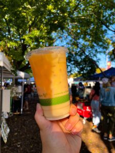 A close-up of a hand holding an iced Pumpkin Up Chai latte in a clear cup with a green compostable print. The latte has a rich orange hue, capturing the essence of fall. In the background, people are strolling through logan square farmers market under the shade of large, leafy trees in October 2023.