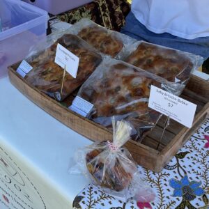 A close-up of a wooden tray displaying several packaged loaves of sourdough focaccia and chocolate baby brioche babka. Each item is labeled with a small sign, showing prices of $11 for the focaccia and $7 for the babka. In the foreground, there is a smaller bagged item, also from 1902 bakery, sitting on a white and floral patterned tablecloth.