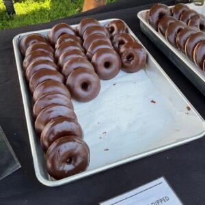 A close-up of a silver bakery tray displaying several rows of chocolate covered, cinnamon sugar coated baked donuts. The chocolate glaze shines on each donut, and the tray has a large empty space showing how many have been sold.