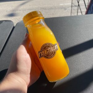 A bottle of fresh pressed juice from Frogtown Juice in the flavor Sonny (mango, pineapple, orange) held over a black table cloth with sun shining.