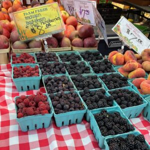 Quart produce boxes of early fall raspberries, black raspberries, and blackberries with a sign reading $8.00, next to donut peaches and in front of apples and peaches.