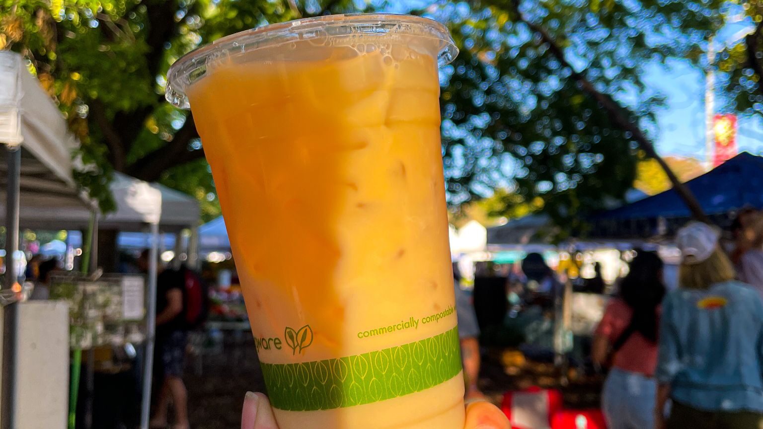 A close-up of a hand holding an iced Pumpkin Up Chai latte in a clear cup with a green compostable print. The latte has a rich orange hue, capturing the essence of fall. In the background, people are strolling through logan square farmers market under the shade of large, leafy trees in October 2023.