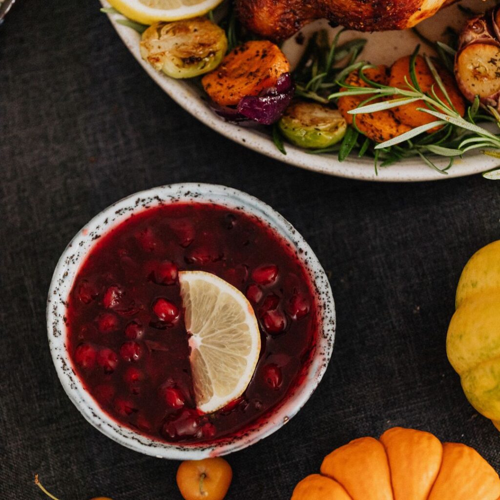 A close-up of a bowl of Holly Berry Cranberry Sauce, rich in color and garnished with a lemon slice. The sauce sits alongside roasted vegetables and small decorative pumpkins, tying in the festive mood.