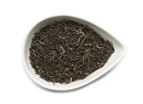 A close-up of loose-leaf Kumaon Black Tea displayed in a white teardrop-shaped dish. The tea leaves are dark brown with occasional golden tips, showcasing their rich, twisted, and slightly curled texture.