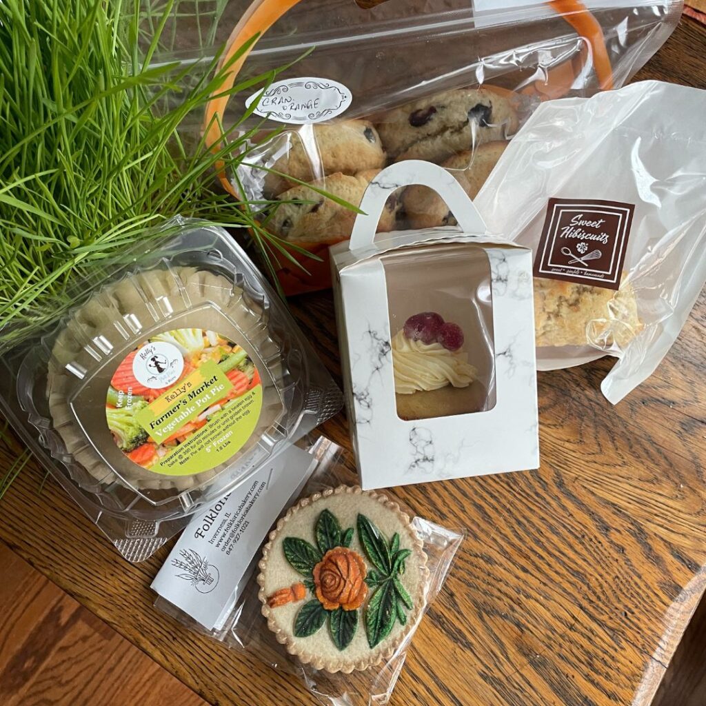 A display of farmers market goodies featuring a small pot of vibrant cat grass, a pack of vegetable pot pies labeled “Kelly’s Farmers Market Vegetable Pot Pie,” a decorative cookie with a painted floral design from Folklorica Bakery, a boxed cupcake with candied cranberry topping, a pack of Sweet Hibiscuits scones, and a bag of cranberry orange scones from Folklorica. The items are arranged on a wooden table.