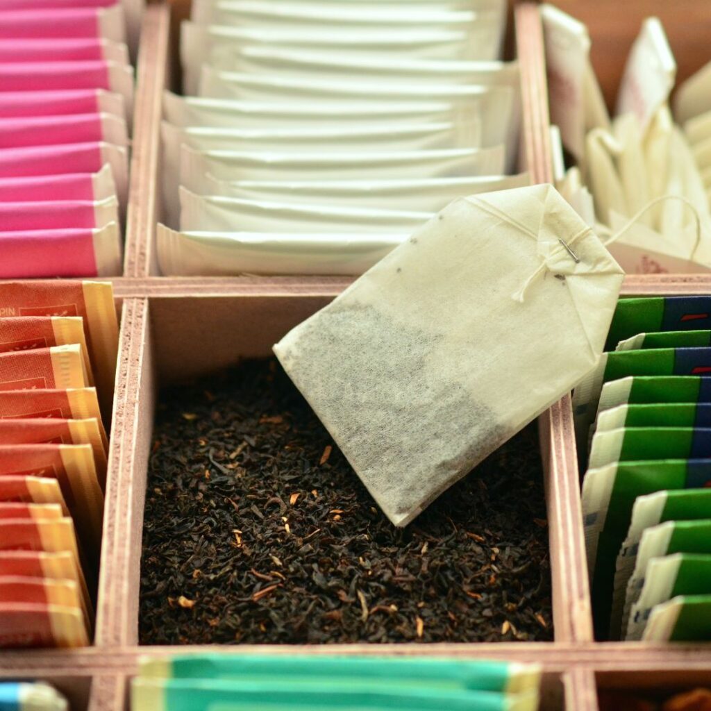 A tea bag filled with tiny tea fannings resting on a wooden box compartment that also contains loose fannings. The small, fine particles of the fannings demonstrate their low quality compared to whole-leaf tea.