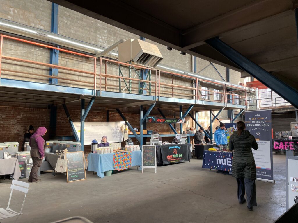 An industrial-style indoor market with exposed brick walls and metal beams. Vendors are set up with tables displaying various goods, and shoppers browse the booths. A second level is visible with additional vendor space.