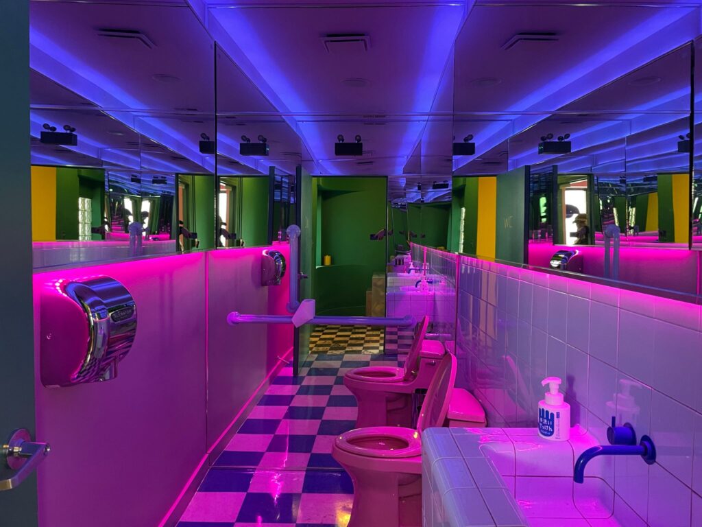 A colorful and modern bathroom featuring vibrant neon lighting in pink and blue. The space includes mirrored walls, tiled floors, and pink toilet fixtures, creating a bold and futuristic look.