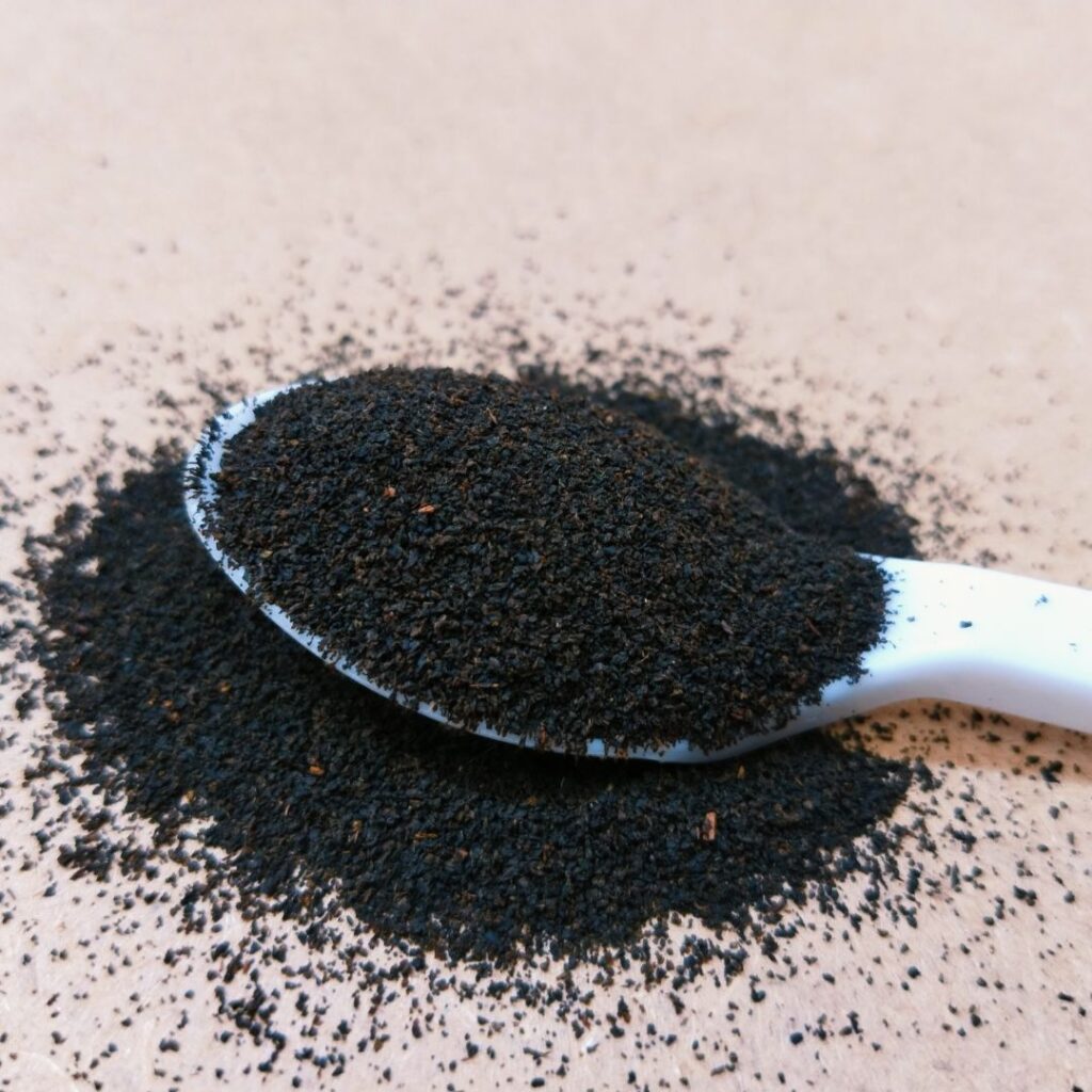 A close-up of a white spoon filled with fine black tea dust (fannings) resting on a pile of scattered tea particles on a beige background. The tiny size of the tea dust highlights its low-grade quality.