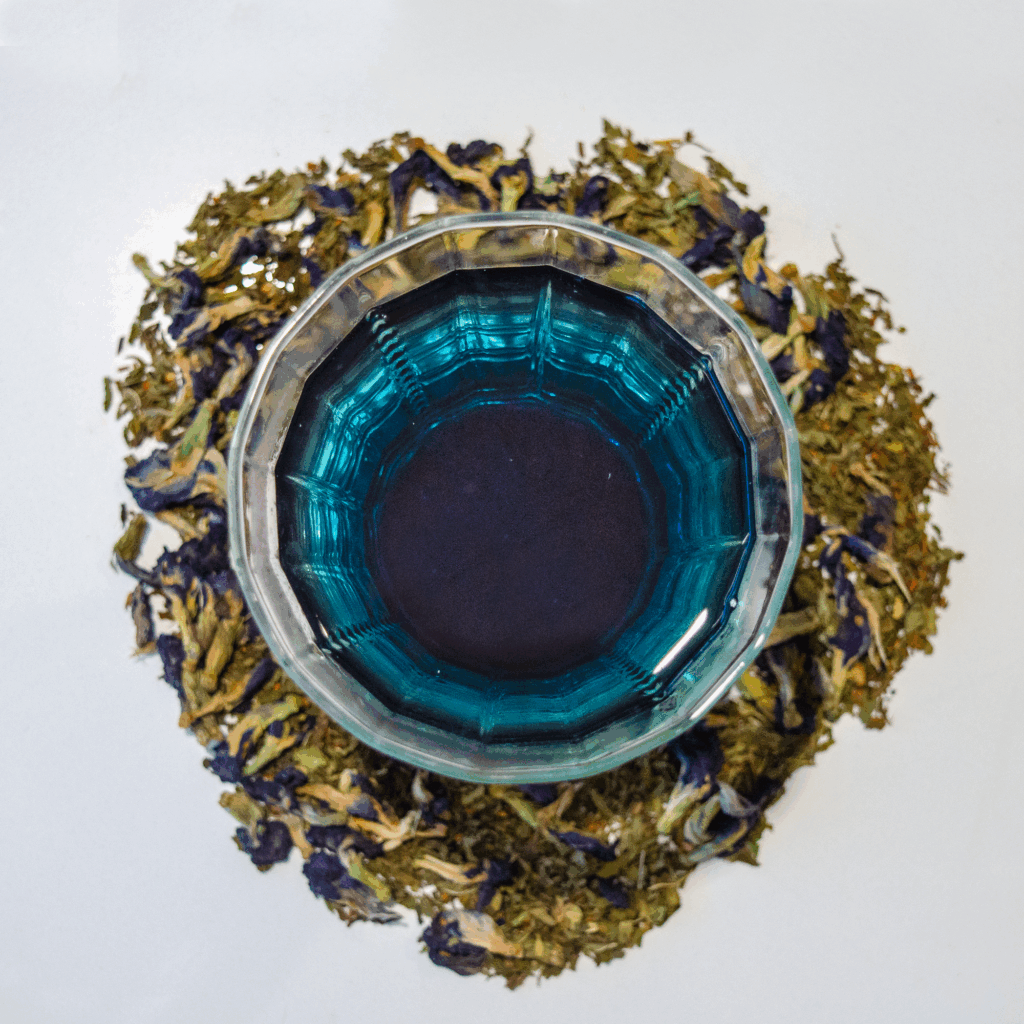 A glass teacup filled with vibrant blue tea sits centered on a white background, surrounded by a circular arrangement of loose leaf rooibos, spearmint, and butterfly pea flowers, infused with vanilla essence.
