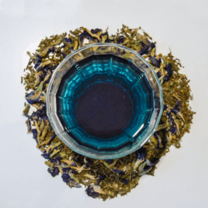 A glass teacup filled with vibrant blue tea sits centered on a white background, surrounded by a circular arrangement of loose leaf rooibos, spearmint, and butterfly pea flowers, infused with vanilla essence.