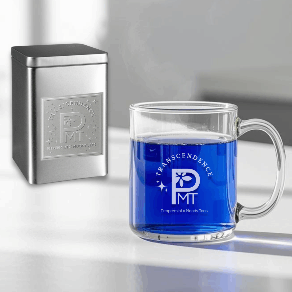 A glass mug of vibrant blue Transcendence tea sits on a white surface beside a silver embossed tin, both featuring the Transcendence logo for the Peppermint x Moody Teas collaboration. Steam rises gently from the mug, highlighting the tea’s striking color and elegant presentation.