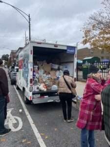 A large U-Haul truck with its back door open, overflowing with bags and boxes of donated food at the Lincoln Park Farmers Market food drive. Volunteers and community members stand nearby on the street, helping load additional items into the truck because it is completely full. The sky is overcast and autumn leaves scatter the ground.